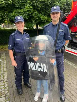 policjant i policjantka z dzieckiem ubranym w kask policyjny i z tarczą policyjną